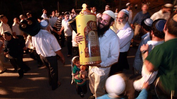 Ein kleines Mädchen hält eine Puppe und ihren Vater, der mit einer Torah-Rolle zwischen jüdischen Siedlern tanzt