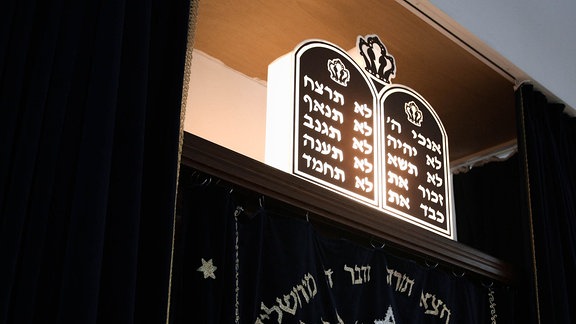 Eine Tafeln mit hebräischer Schrift über einem Holzschrank, der von einem blauen Samtvorhang verdeckt wird