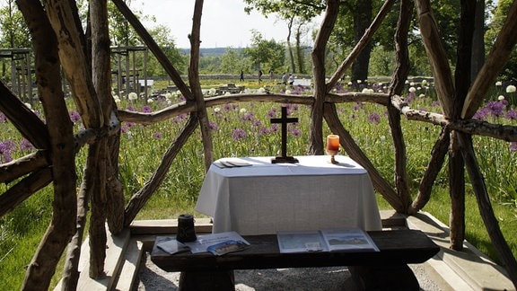 Altar im Freien unter zusammengestzten Stämmen. Im Hnitergrund ist ein Garten mit Blumen und Kräutern