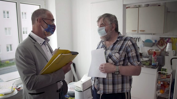 Zwei Männer stehen mit Mund-Nasen-Maske in einer Wohnung.