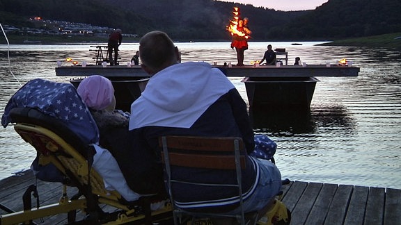 Auf einer schwimmenden Plattform auf einem See steht eine Frau und vollführt Kunststücke mit Feuer. Am Ufer sitzen ein Kind und ein junger Mann und schauen zu.