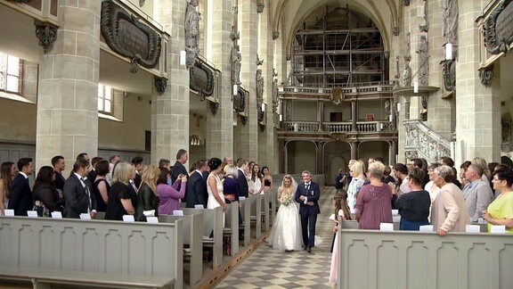 Eine Frau im Brautkleid wird von einem Mann durch den Kirchgang zum Altar geleitet. Die Hochzeitsgäste stehen und schauen zur Braut.