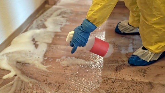 Reportage Tatortreiniger: Eine Hand in Schutzkleidung sprüht Reinigungsmittel auf einen Fußboden.
