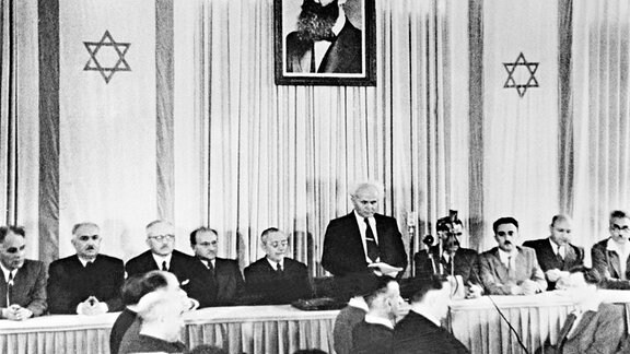 Der erste israelische Premierminister David Ben-Gurion (stehend) verkündet in Tel Aviv vor Mitgliedern der jüdischen Ratsversammlung die Gründung des Staates Israel, während über ihm ein Porträt von Theodor Herzl, Begründer des politischen Zionismus, hängt (Archivfoto vom 14. Mai 1948).