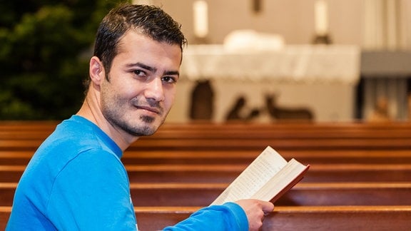 Ein junger Mann sitzt in einer Kirche und schaut in die Kamera.
