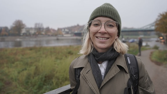 Eine Frau mit Mütze und Brille lächelt in die Kamera, hinter ihr sieht man das Flussufer der Elbe in Dresden.