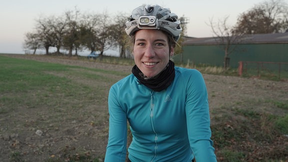 Eine Radsportlerin lächelt beim Traningsstopp auf einer Landstraße in die Kamera.