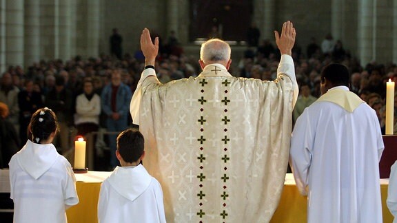 Priester steht mit erhobenen Händen zu den Kirchenbesuchern gewandt. Neben ihm stehen Messdiener.
