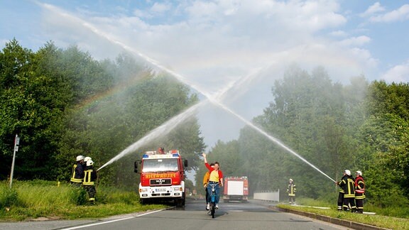 Eine Frau und ein Mann fahren mit ihrem Tandem durch Wasserfontänen der Feuerwehr, darüber strahlt ein Regenbogen.