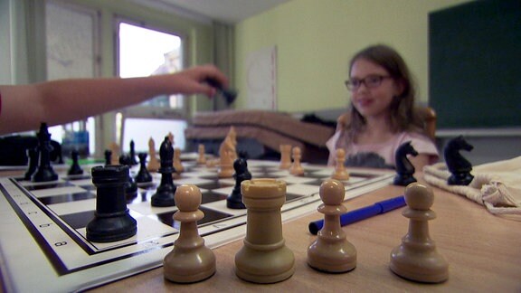 Schachfiguren vor einem Schachbrett an dem zwei junge Mädchen spielen.