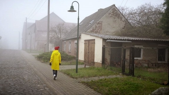 Frau im gelben Regenmantel auf Dorfstraße bei Nebel unterwegs.