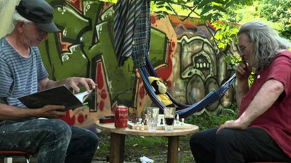 Filmszene - zwei Männer sitzen an einem Tisch im Garten.