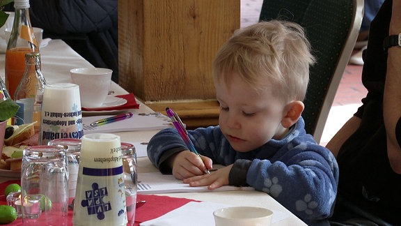 Ein kleiner Junge malt mit einem Stift auf einen Zeichenblock während er an einem Tisch sitzt.