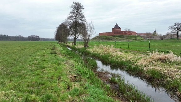 Ein kleiner Fluss, im Hintergrund eine rote Burg.