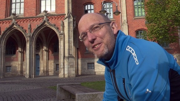 Ein Mann mit kurzen Haaren, Brille und blauer Sportjacke, sitzt vor einem Schulgebäude und schaut lächelnd in die Kamera.