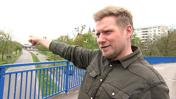 Ein Mann steht auf einer Brücke und zeigt mit seinem rechten Arm auf einen Punkt im Hintergrund.