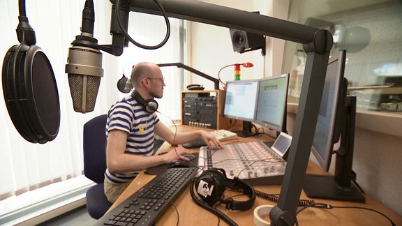 Ein Mann arbeitet im Hintergrund am Computer, vor ihm ein großes Mischpult, im Vordergrund hängen zwei Mikrofone.