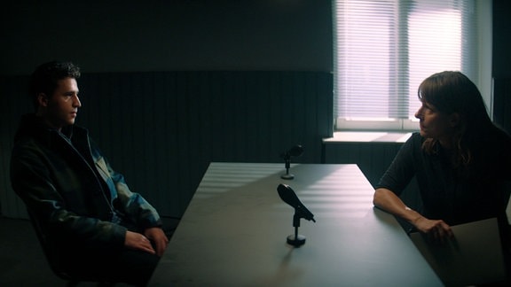 Filmszene - Komissarin und Verdächtiger sitzen sich beim Verhör in einem abgedunkeltem Raum gegenüber. Etwas Licht fällt im Hintergrund durch die Jalousie eines Fensters.