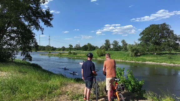 Zwei Männer stehen mit ihren Fahrrädern am Ufer der Saale und beobachten vorbeikommende Padelboote.
