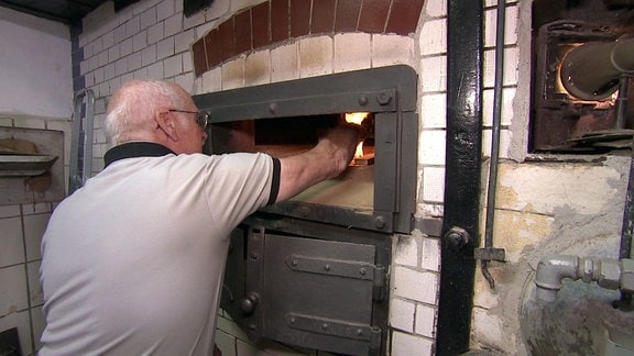 Ein Mann legt ein Holzscheid in einen Ofen um das Feuer weiter anzuheizen.