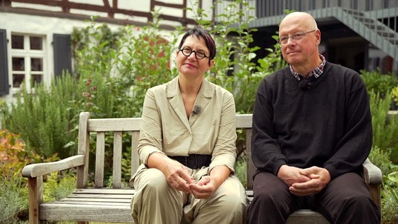 Zwei Personen sitzen auf einer Gartenbank und schauen zur Kamera.