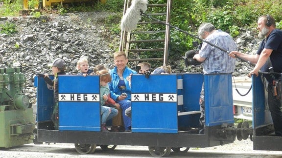 Kinder sitzen in mehreren blauen Wagen einer alten Bergbahn, daneben steht ein Kamerateam, das mit einer Tonangel die Situation einfängt