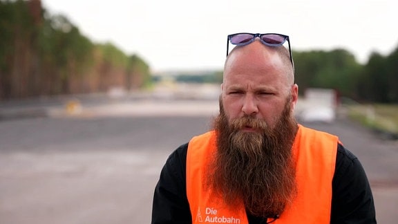 Ein bärtiger Mann mit orangefarbener Warnweste steht auf einer Autobahn.