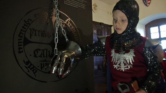 Ein Junge in Ritterrüstung probiert in einer Ausstellung einen echten Ritterhandschuh aus.