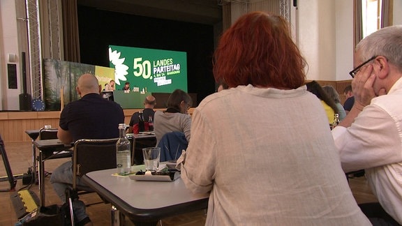 Personen sitzen beim Landesparteitag von Bündnis 90 die Grünen auf und vor einer Bühne.