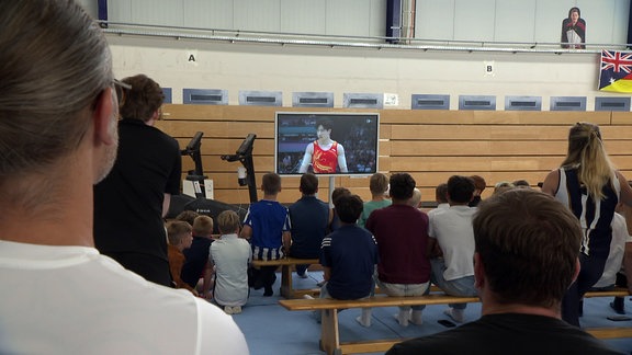 In einer Turnhalle schauen Menschen auf einen Fernseher auf dem Olympia gezeigt wird.