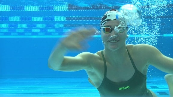 Eine Schwimmerin winkt unter Wasser in die Kamera.