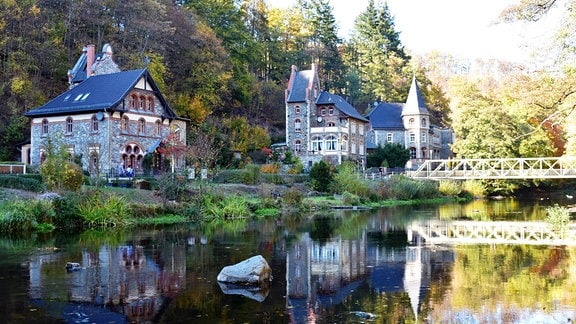 Mehrere steinerne Häuser mit Türmen stehen am Ufer eines Gewässers, auf der rechten Seite ist eine Brücke zu sehen, die Szenerie spiegelt sich im Wasser. 