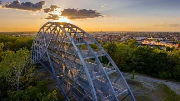 Der Bitterfelder Bogen: ein begehbarer Bogen mit vielen Querstreben aus Metall, die Sonne geht im Hintergrund unter.