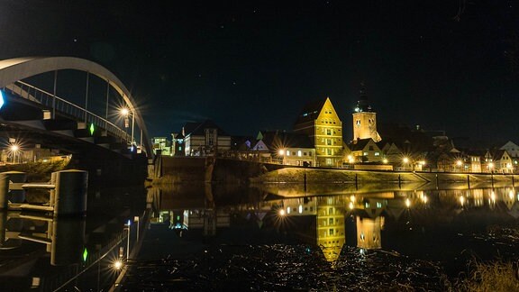 Eine Stadt bei Nacht: beleuchtete Häuser und eine Brücke, die sich im Fluss spiegeln.