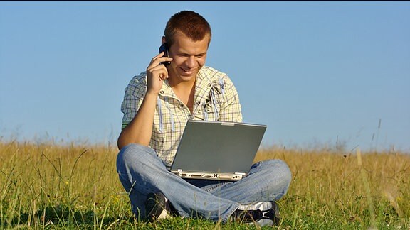 Ein junger Mann sitzt mit einem Laptop und einem Telefon am Ohr auf einer Wiese