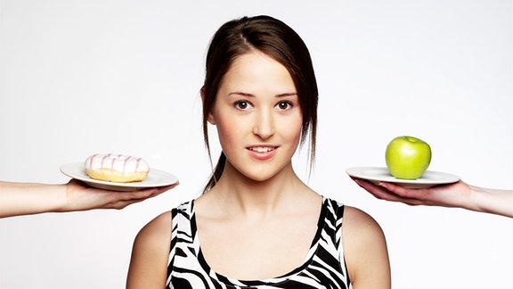 Symbolbild Diät: eine junge Frau, hin und her gerissen zwischen einem Donut und einem Apfel.