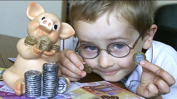 Ein Junge mit Münzen und Geldscheinen, neben ihm ein Sparschwein