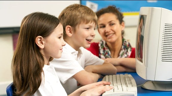 Symbolbild: IT-Unterricht an Schulen - Ein Mädchen und ein Junge werden von einer Lehrerin an einem Computer betreut.