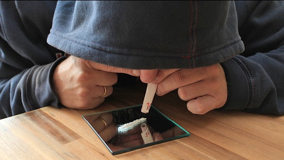 Ein Mann in Kapuzen-Shirt simuliert mit einem aufgerollten 10-Euro-Schein und weißem Pulver auf einem Spiegel das Konsumieren von Drogen