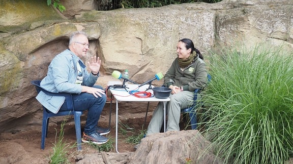 Ein Mann und eine Frau sitzen neben einem Felsen an einem Tisch mit Mikrofonen.