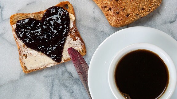 Eine Tasse schwarzer Kaffee und ein Toast mit Marmelade in Herzform stehen auf einem Frühstückstisch