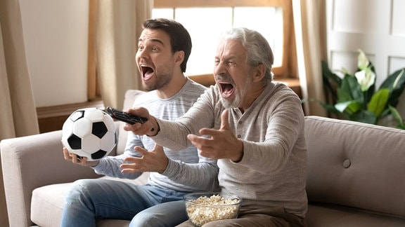 Zwei Männer sitzen mit Fußball und Fernbedienung in den Händen auf einem Sofa und jubeln