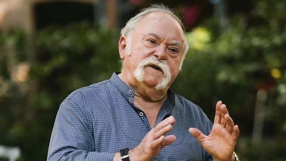 Ein als der Kabarettist Gunter Böhnke bekannter Mann mit schütterem grauen Haar und markantem grauen Schnauzbart steht in einem Garten, spricht und gestikuliert mit den Händen