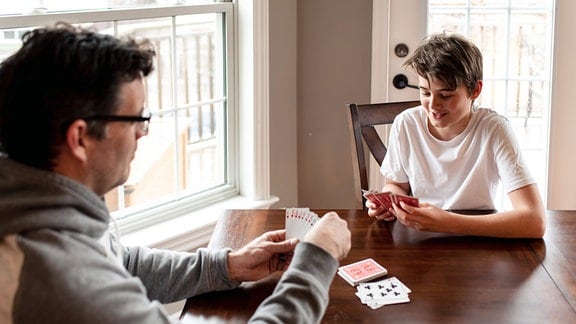 Heranwachsender Junge und sein Vater spielen gemeinsam Karten am Tisch. 
