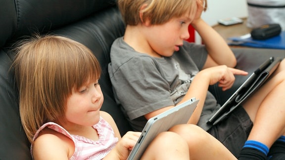 Zwei Kinder im Vorschulalter spielen auf einer Couch sitzend mit ihren Tabletts