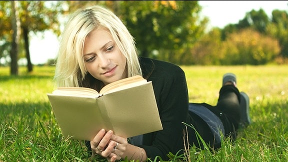 Eine junge blonde Frau liegt in einem Buch lesend auf einer Wiese