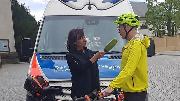 Eine Frau mit Einem Mikrofon steht vor einem Mann mit Fahrrad