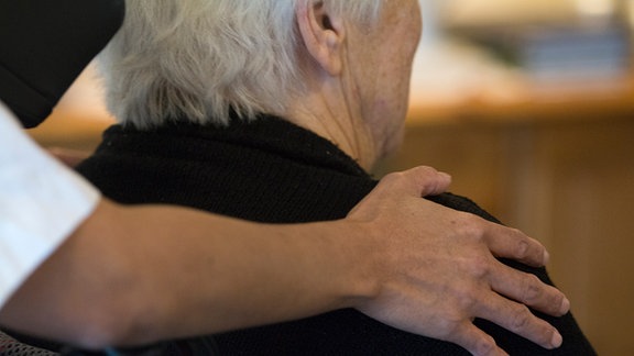 Eine Pflegekraft legt die Hand auf die Schulter einer älteren Frau auf.
