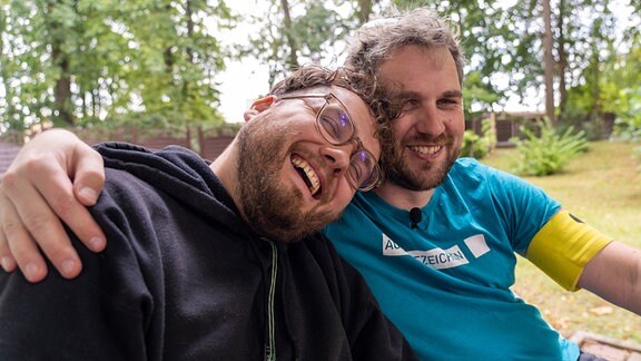 Konstantin Henß und Daniel Martin (v.l.n.r.) umarmen sich und lachen dabei.