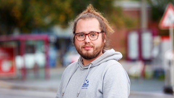 Ein Portrait von Konstantin Henß. Er hat seine langen Haare zu einem Zopf gebunden und blickt direkt in die Kamera. Konstantin trägt einen grauen Kapuzenpullover und eine orange-braune Brille.